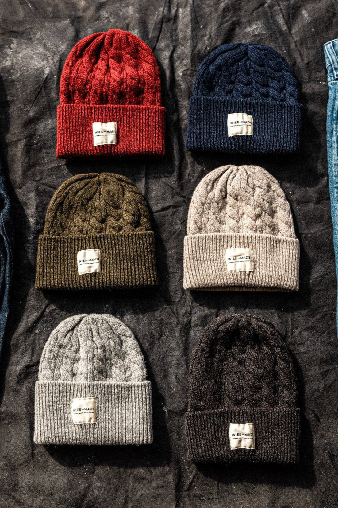 Six cable-knit wool beanies in red, navy, olive, oatmeal, gray, and black laid flat on dark canvas.