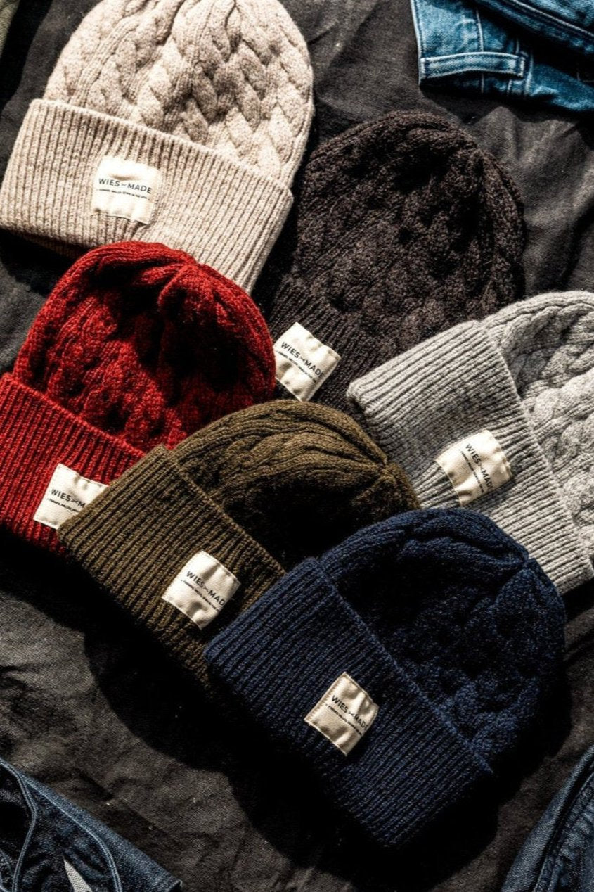 Cable-knit wool beanies in cream, red, brown, olive, gray, and navy arranged on denim.