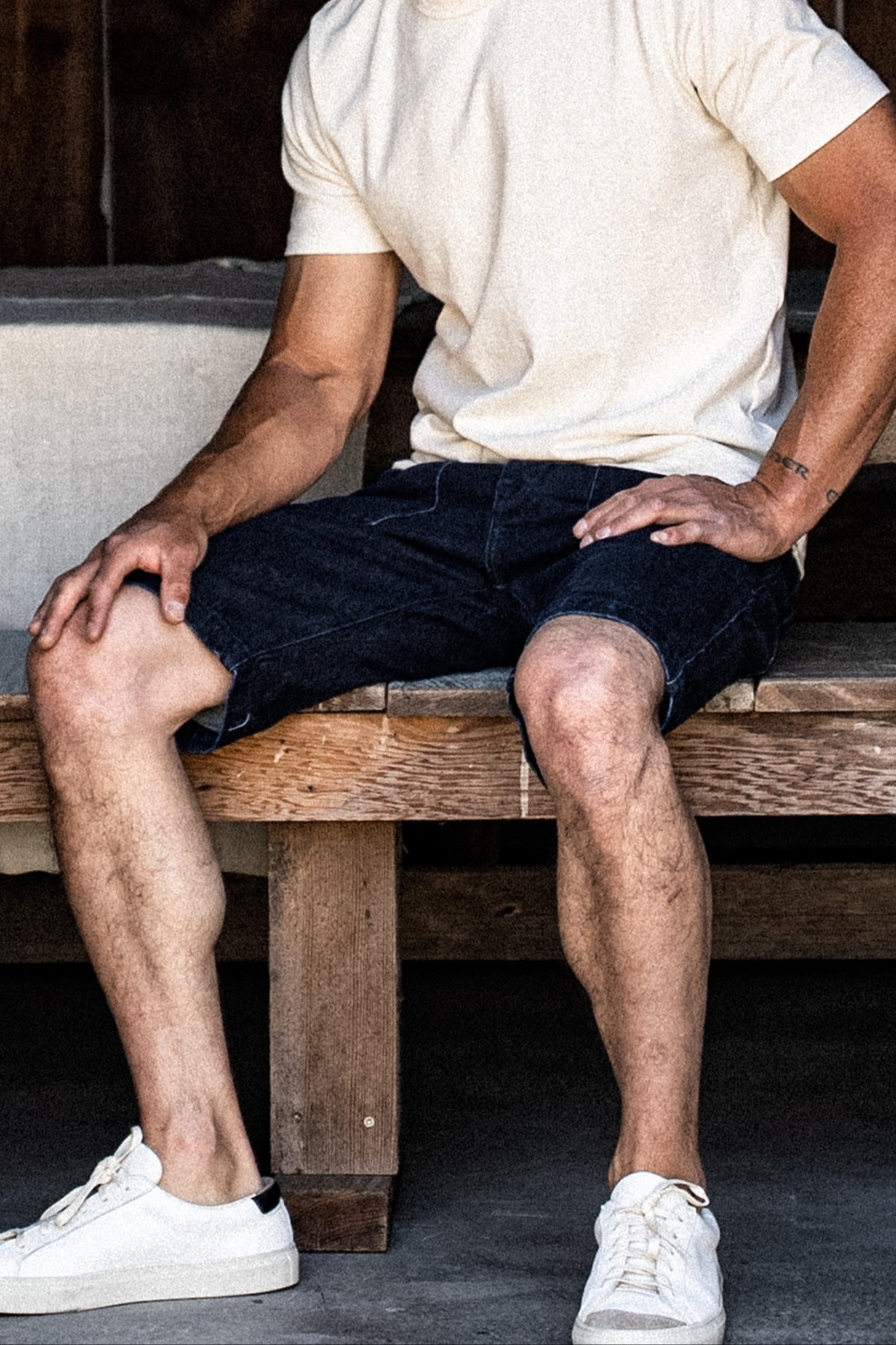 Man seated on a wooden bench wearing dark ocean wash denim military shorts, cream tee, and white sneakers.