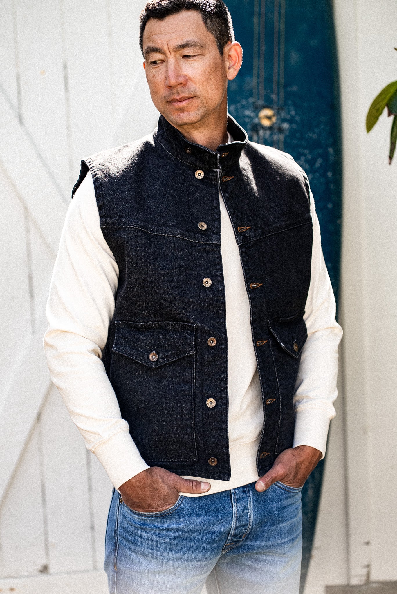 Man wearing black broken twill denim James Vest over cream sweater outdoors.