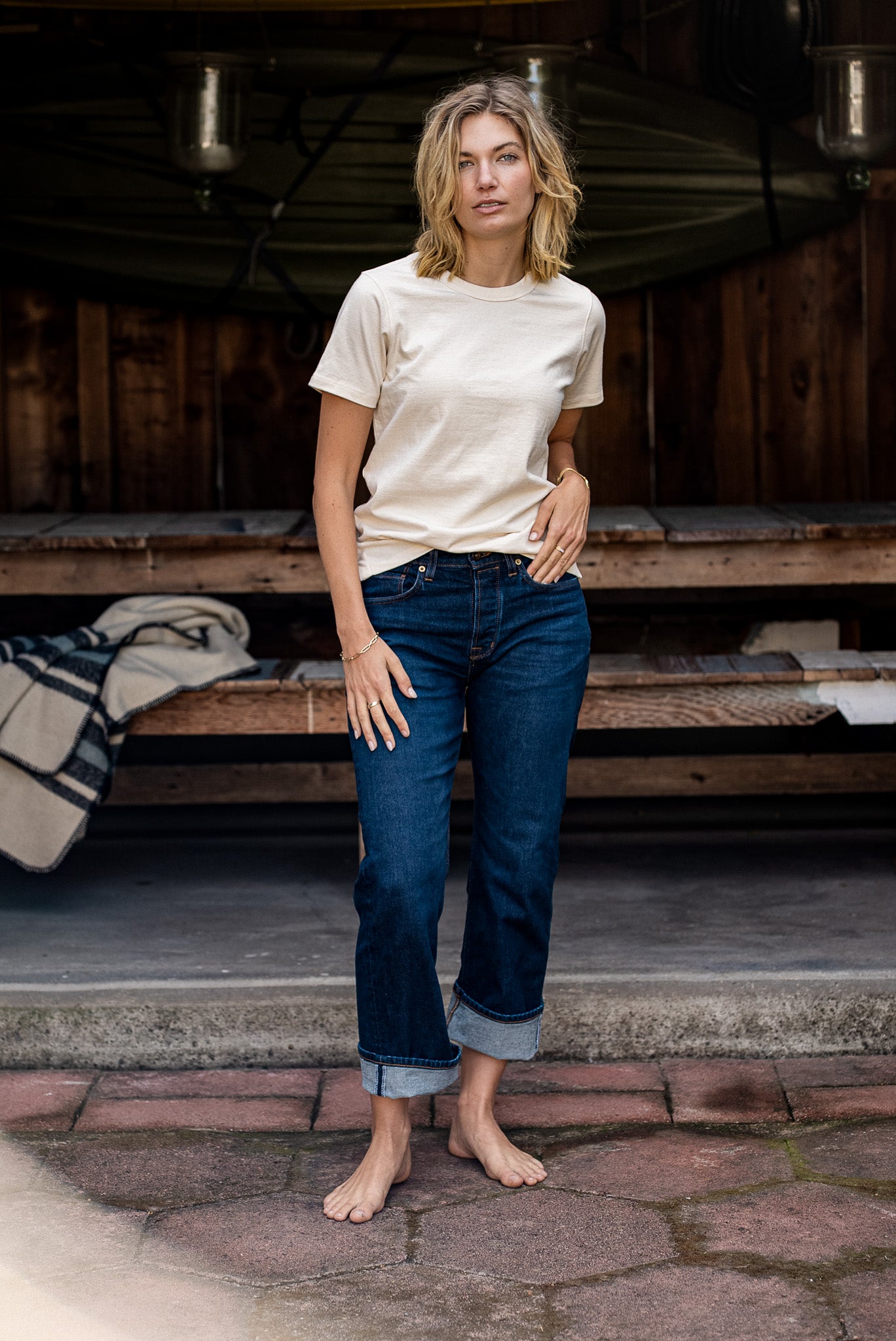 Woman wearing the natural Reyes boxy tee with cuffed dark denim jeans outside a wooden cabin.