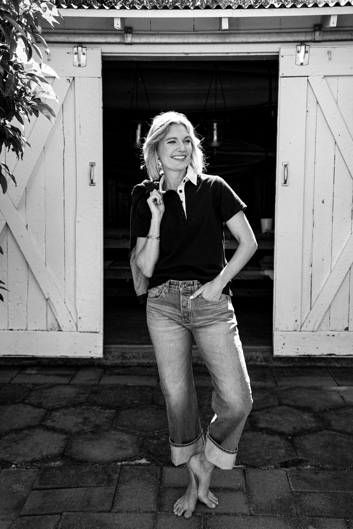 Woman in a short-sleeve dark rugby jersey with contrast collar and cuffed jeans, standing in front of open barn doors.