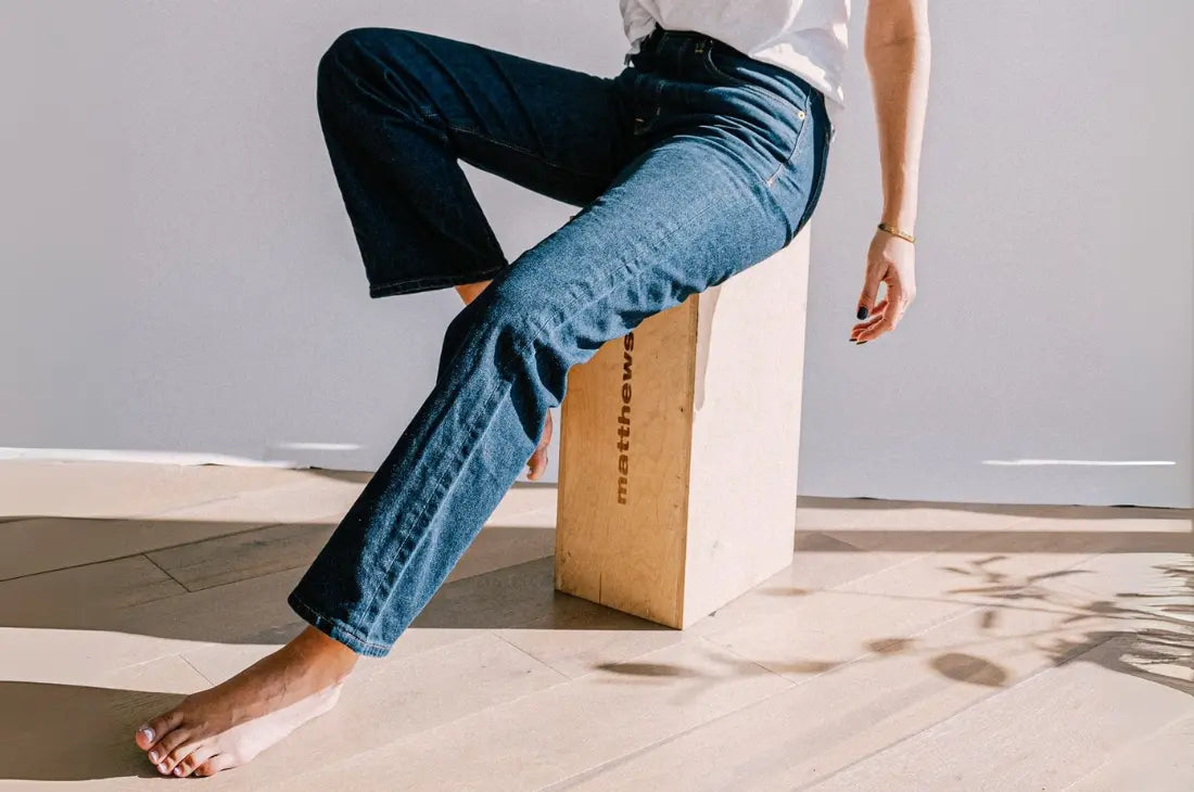 Straight leg jeans in dark blue denim worn while sitting on a wooden block.