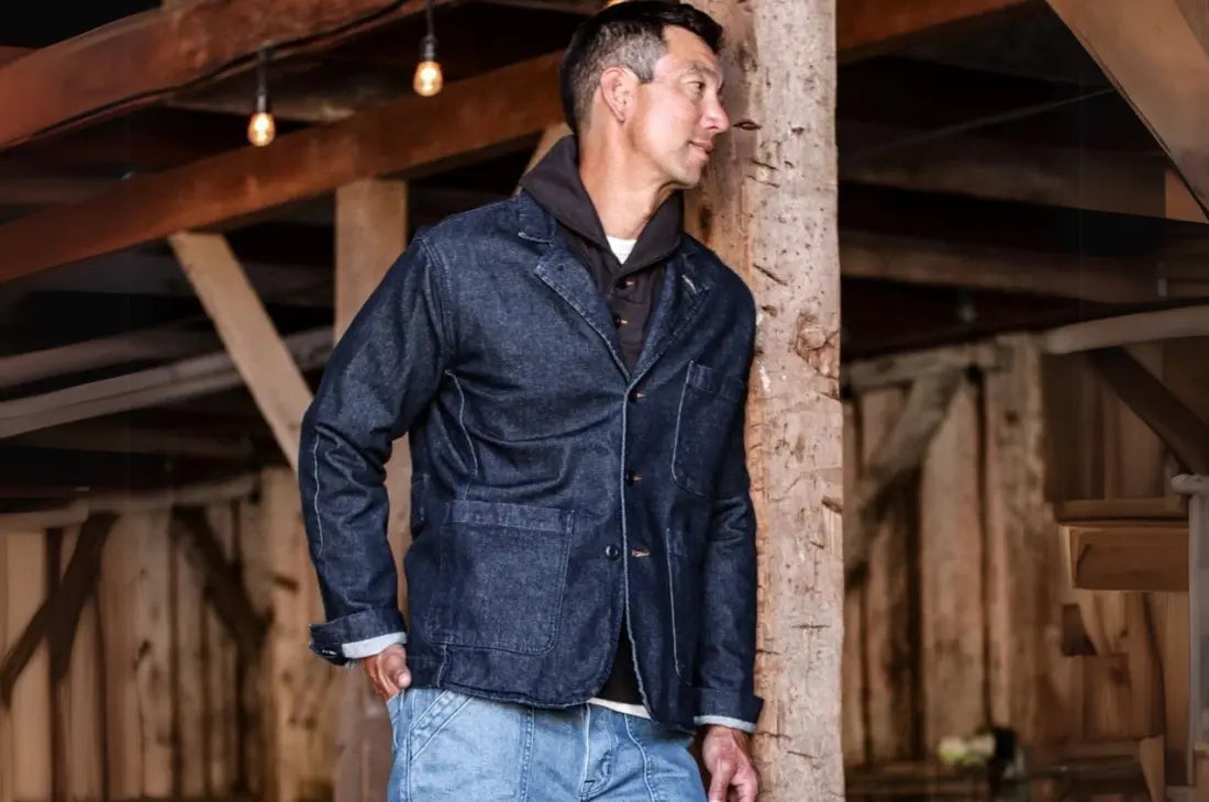 Heritage denim jeans styled with a dark jacket and layered shirt in a rustic barn setting.