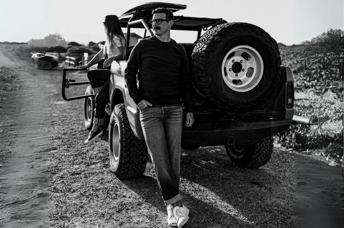 Raw denim jeans worn by man leaning against off-road vehicle in rugged outdoor setting with black and white filter