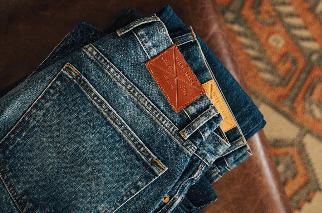 Unwashed denim jeans stacked on a leather surface, featuring contrast stitching and leather brand patches