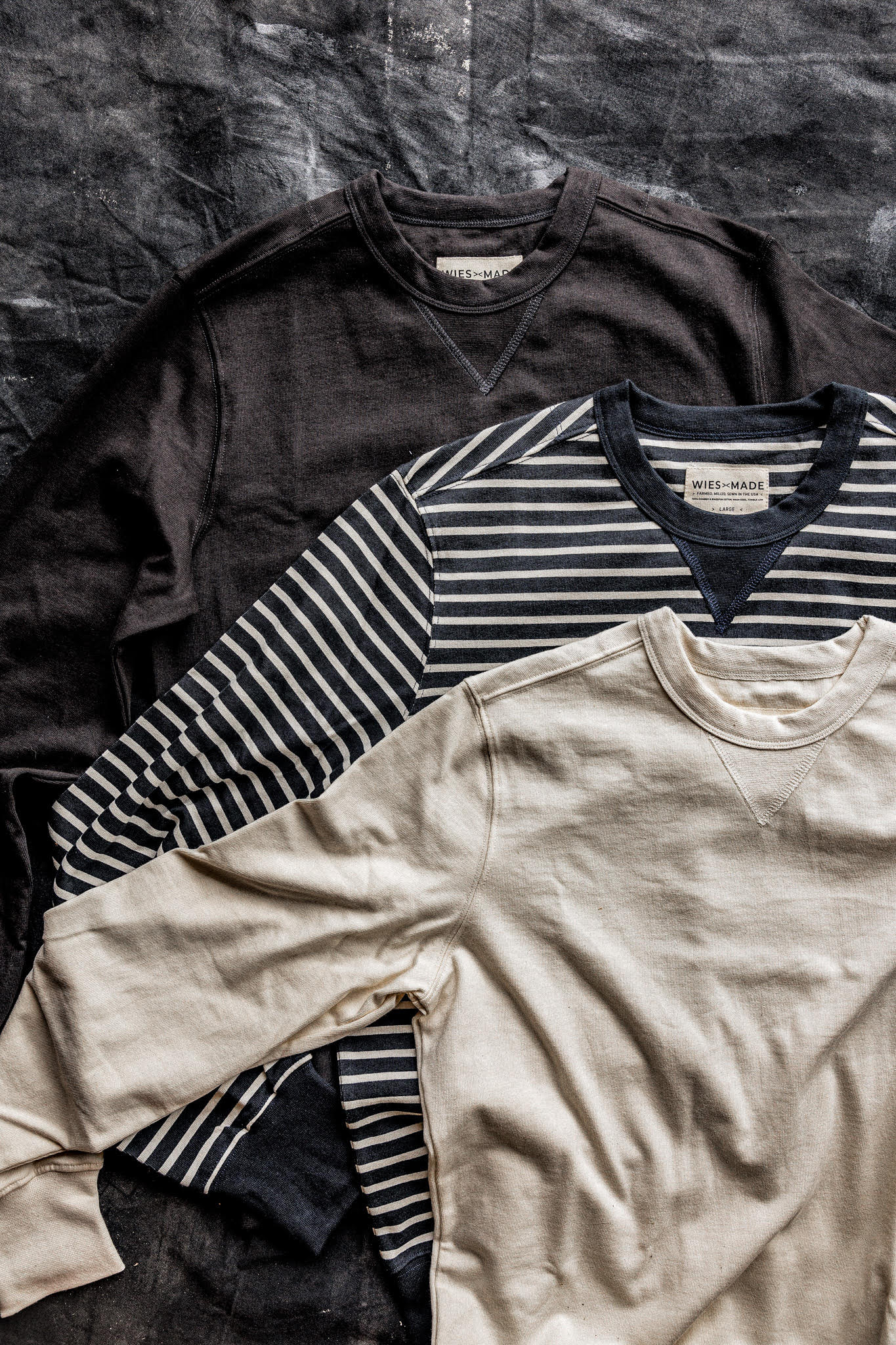 Three Stinson rugby jersey pullovers in basalt, navy stripe, and natural cream laid flat on a dark canvas.