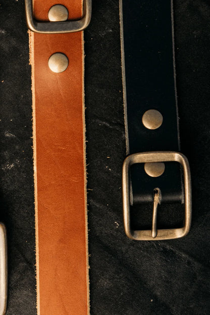 Close-up of saddle tan and black latigo leather belts with solid brass buckles and rivets.
