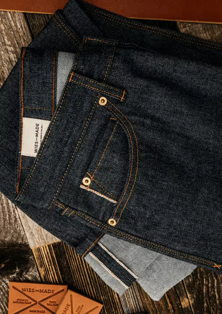Men’s selvedge denim jeans folded on a wooden surface, showcasing premium dark indigo fabric, contrast stitching, and signature edge detailing for a refined artisanal look