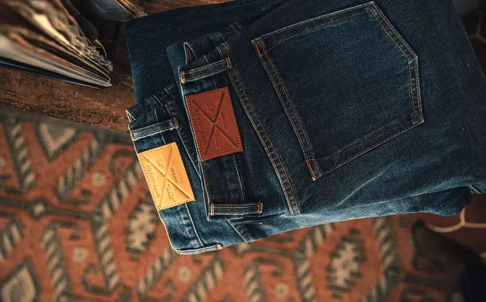 Jack Fit Jeans in Washed Selvedge Denim with a straight cut, neatly folded on a rustic wooden table, featuring a patterned rug and stacked books in the background