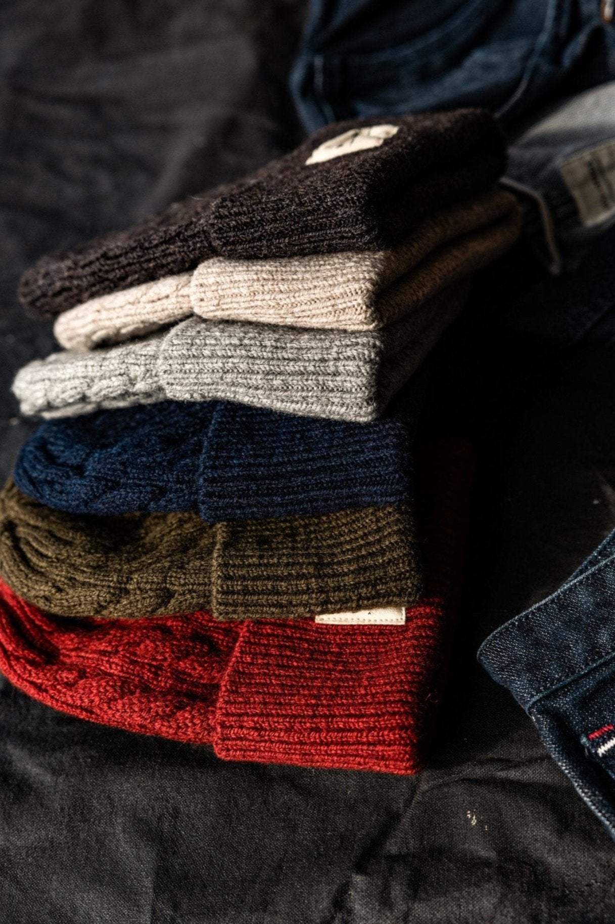 Stack of cable knit wool beanies in brown, oatmeal, gray, navy, olive, and red beside folded denim.