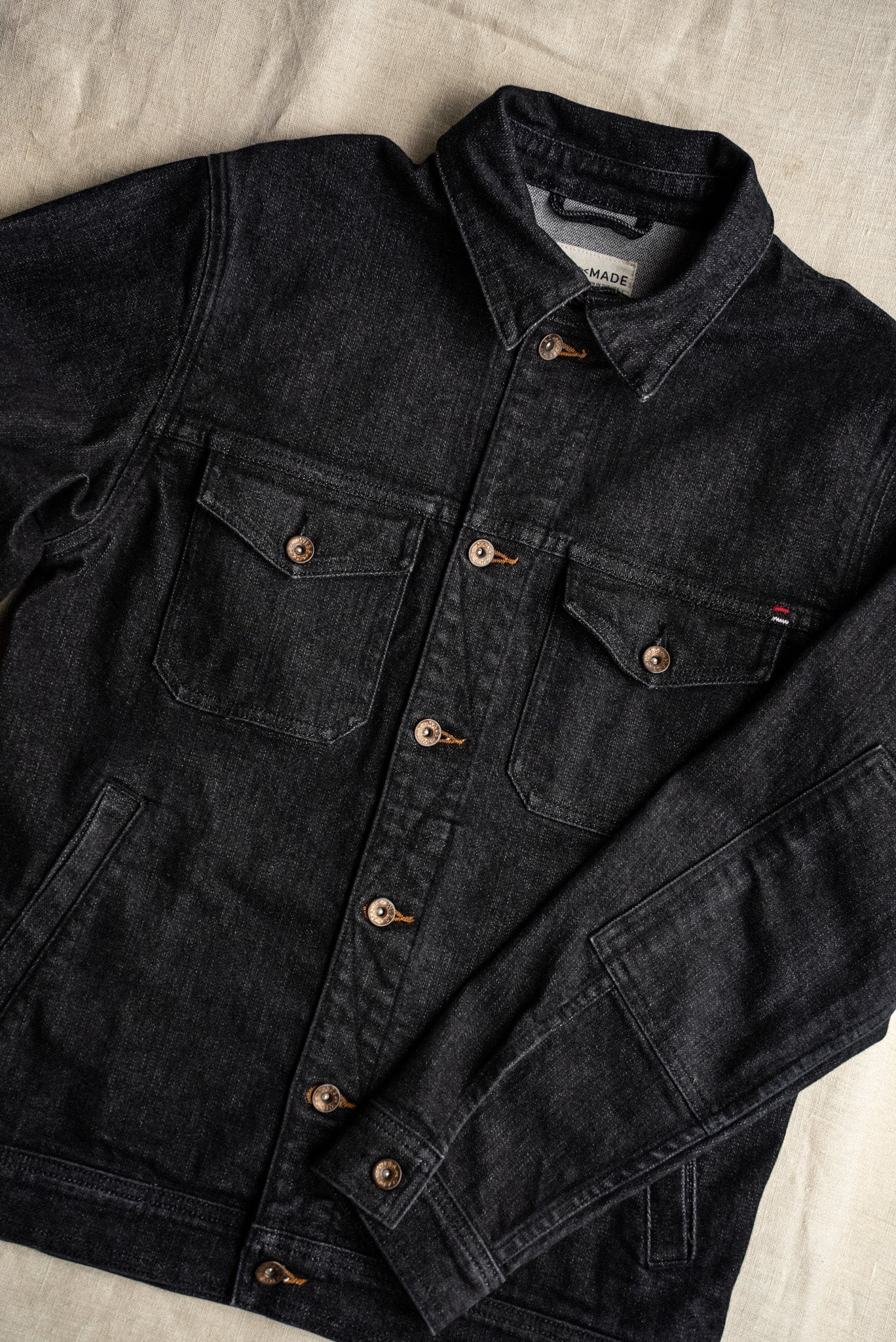 Black broken twill denim jacket with chest flap pockets and brass button closures laid flat on linen.