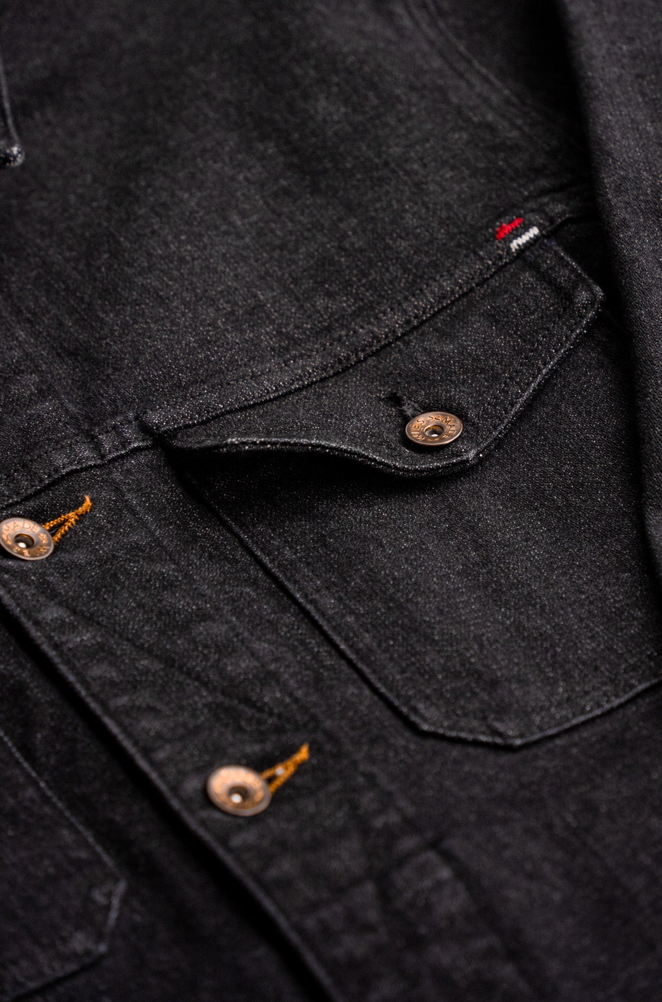 Close-up of black broken twill denim jacket showing chest pocket flap, brass snap and tobacco keyhole buttons.