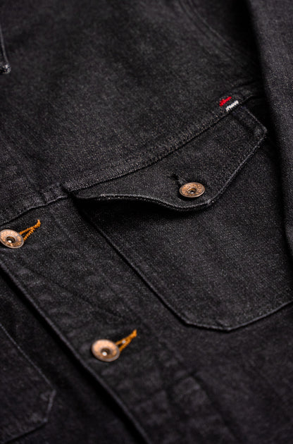 Close-up of black broken twill denim jacket showing chest pocket flap, brass snap and tobacco keyhole buttons.