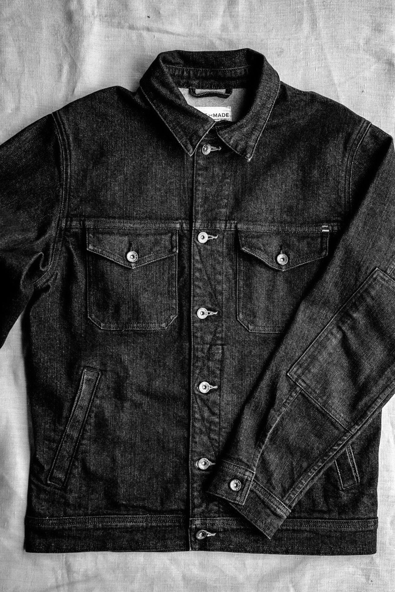 Black broken twill denim jacket flat-laid, showing brass buttons and chest flap pockets.