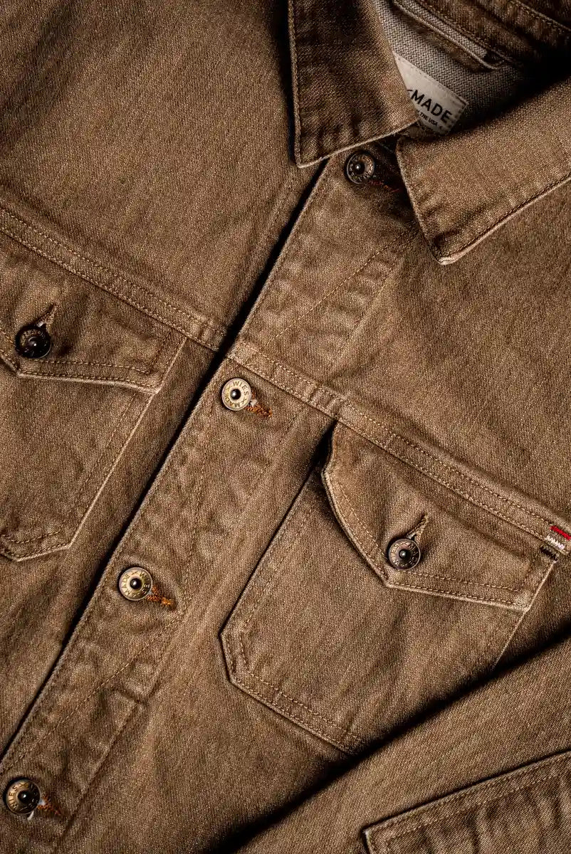 Close-up of wheat-colored broken twill denim jacket showing brass buttons, chest pockets with flaps, and flat felled seams.
