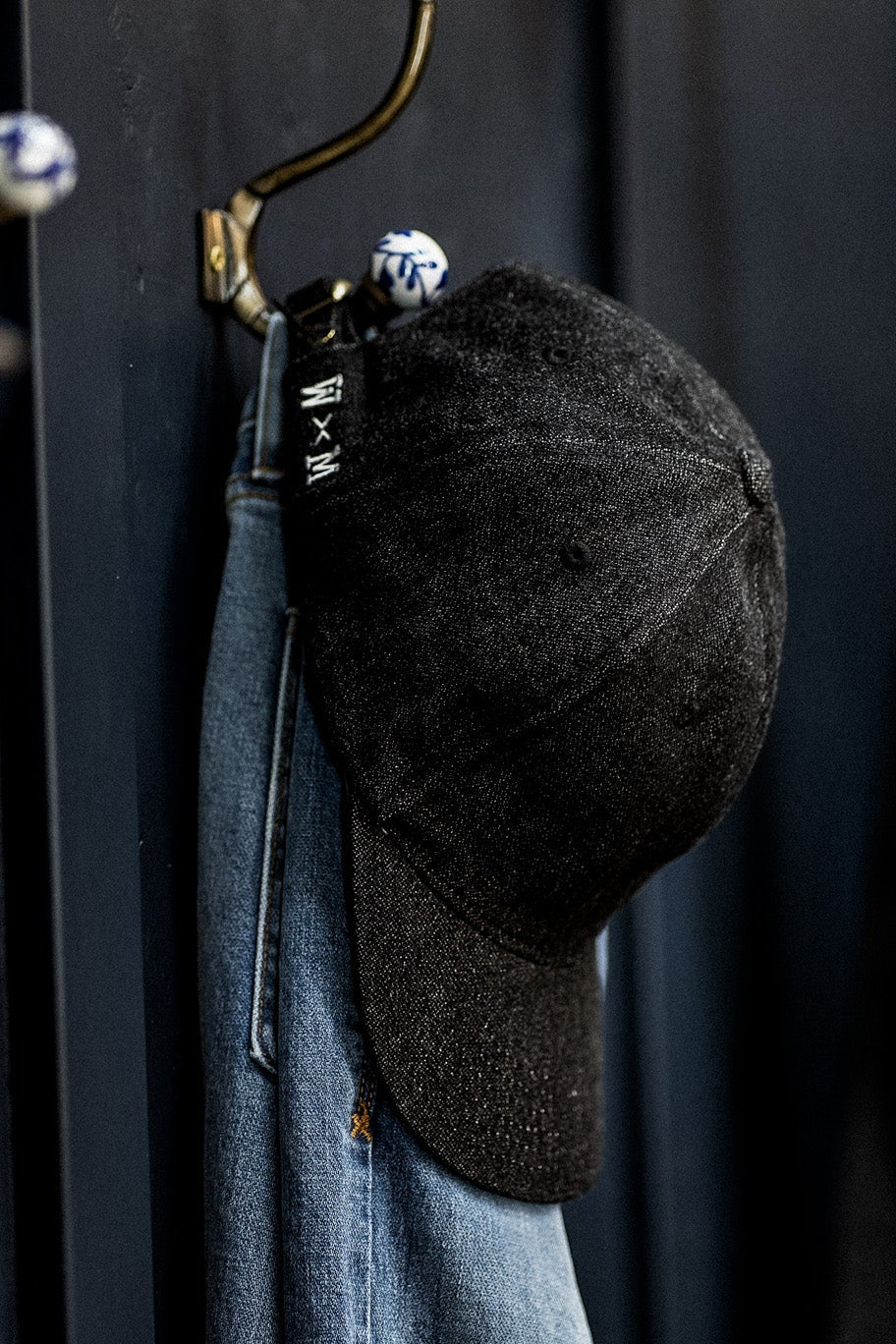 WiesMade black denim ball cap hanging on a brass wall hook next to faded jeans.