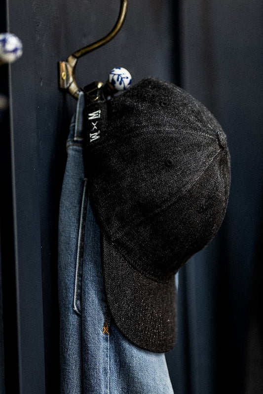 WiesMade black denim ball cap hanging on a brass wall hook next to faded jeans.