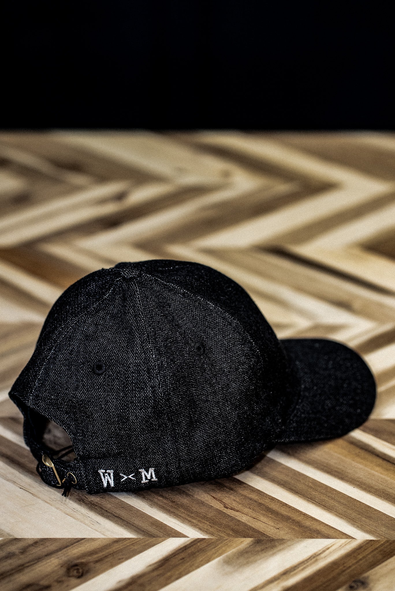 Back view of a WiesMade black denim ball cap with W×M leather buckle strap on herringbone wood.