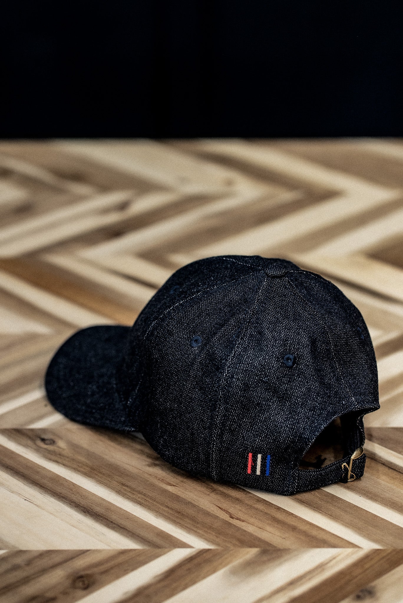 WiesMade black denim ball cap side view showing tricolor label and adjustable strap on herringbone wood.