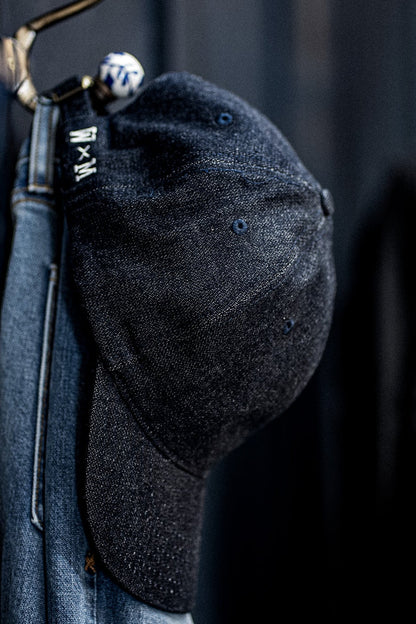Close-up of a WiesMade indigo denim ball cap hanging on a brass hook with the W×M logo strap visible.