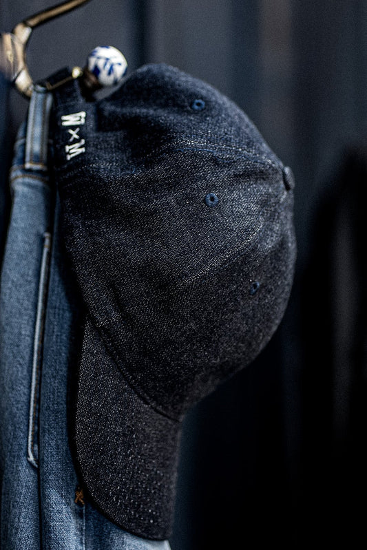 Close-up of a WiesMade indigo denim ball cap hanging on a brass hook with the W×M logo strap visible.