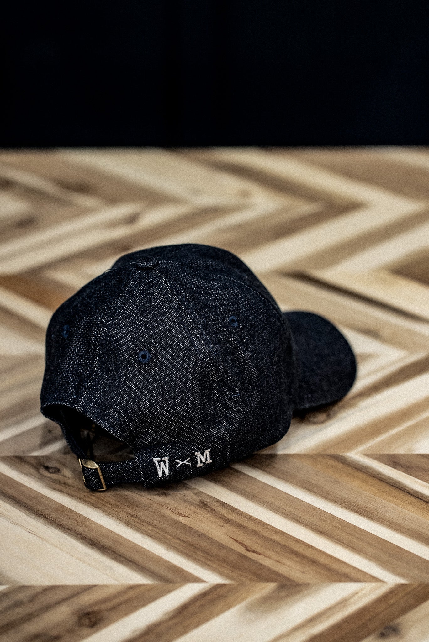 Back view of a WiesMade indigo denim ball cap with W×M leather buckle strap on herringbone wood.