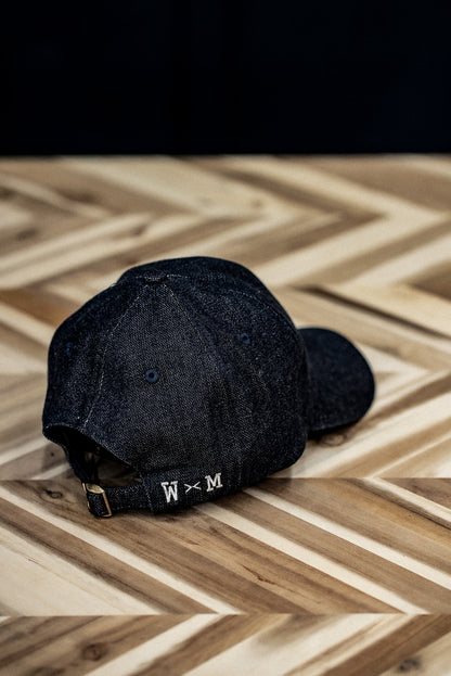 Back view of a WiesMade indigo denim ball cap with W×M leather buckle strap on herringbone wood.
