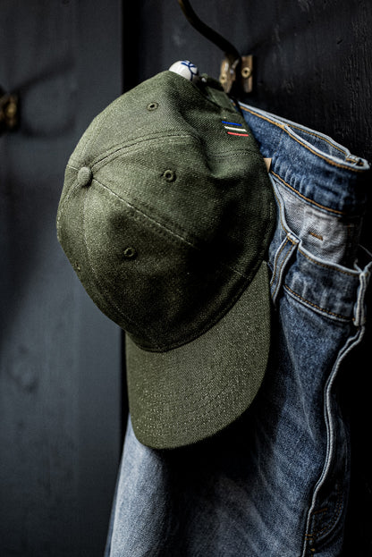 WiesMade olive green ball cap hanging on a brass wall hook alongside faded denim jeans.