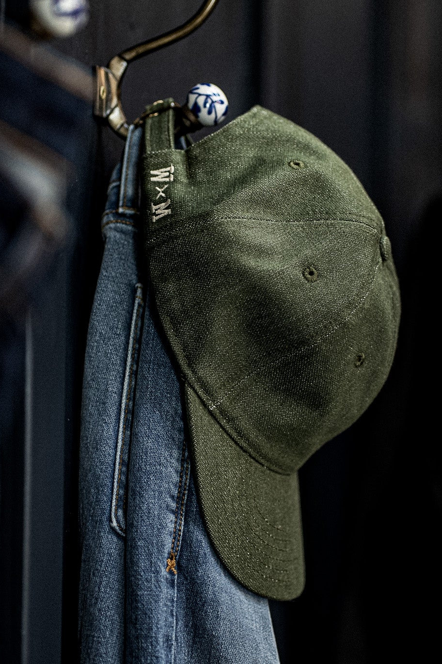 Close-up of a WiesMade olive green ball cap with W×M strap hanging on a hook beside denim jeans.