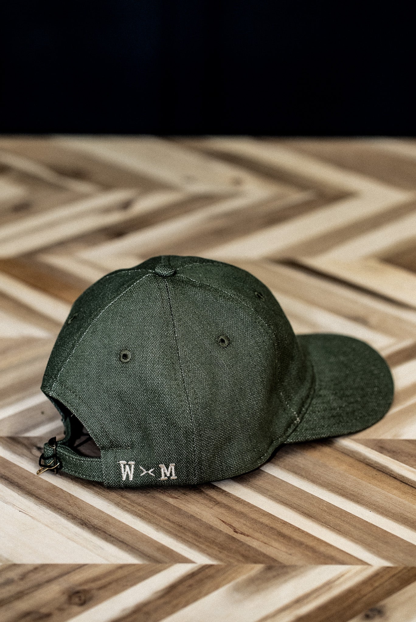 Back view of a WiesMade olive green ball cap with W×M leather buckle strap on herringbone wood.