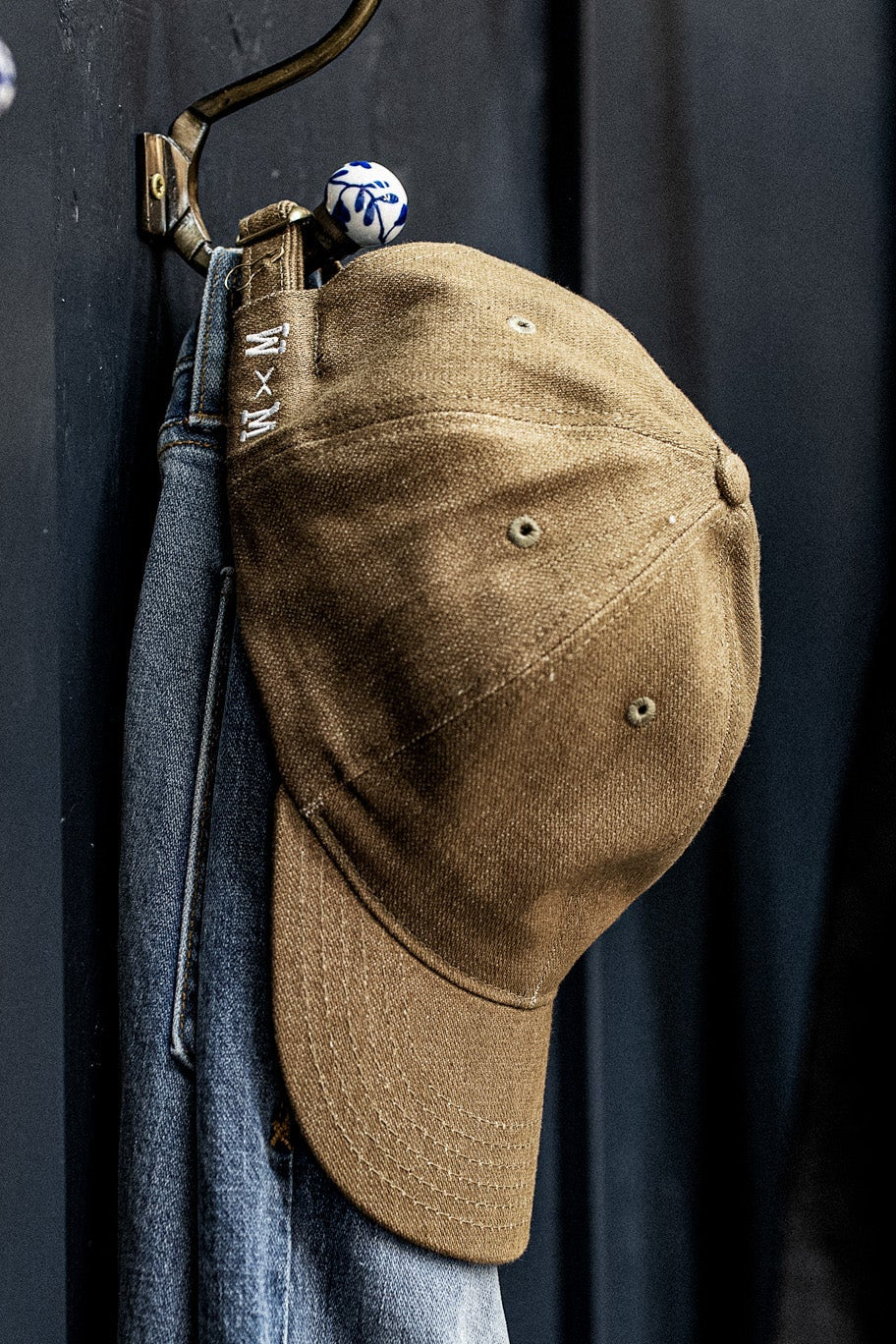 WiesMade wheat canvas ball cap with W×M strap hanging on a brass hook beside denim jeans.