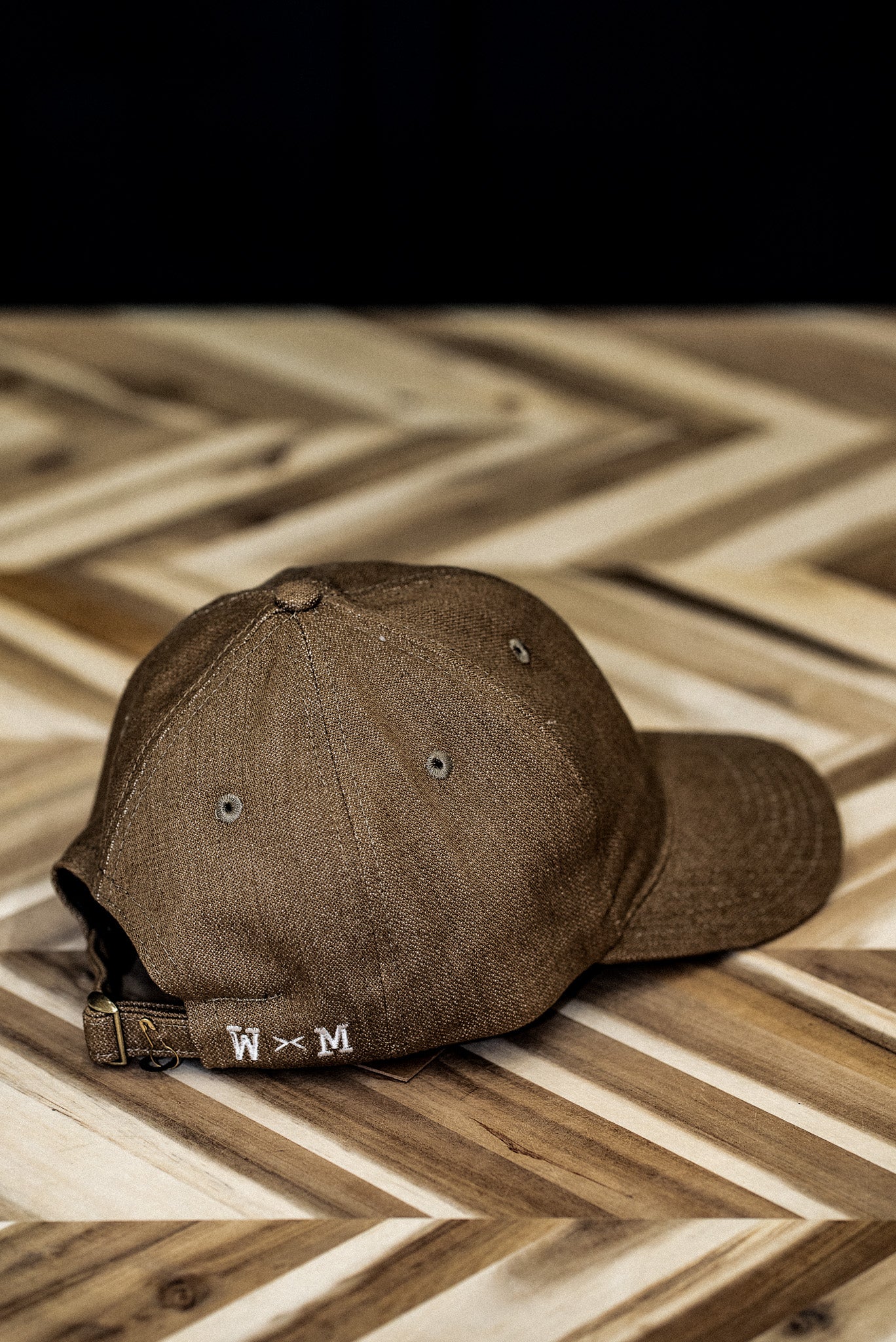 Back view of a WiesMade wheat canvas ball cap with W×M leather buckle strap on herringbone wood.