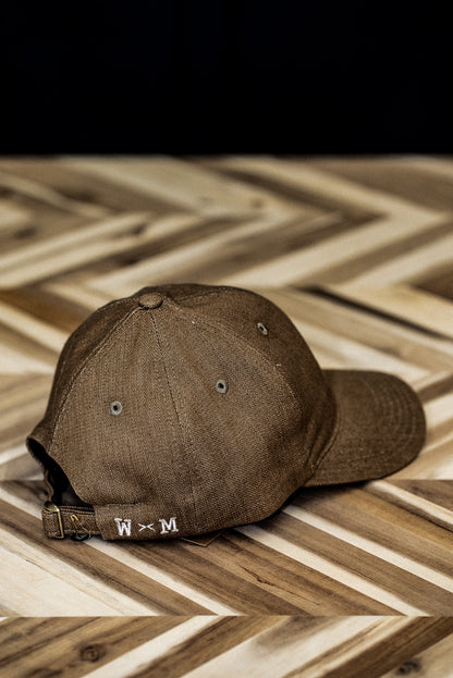Back view of a WiesMade wheat canvas ball cap with W×M leather buckle strap on herringbone wood.