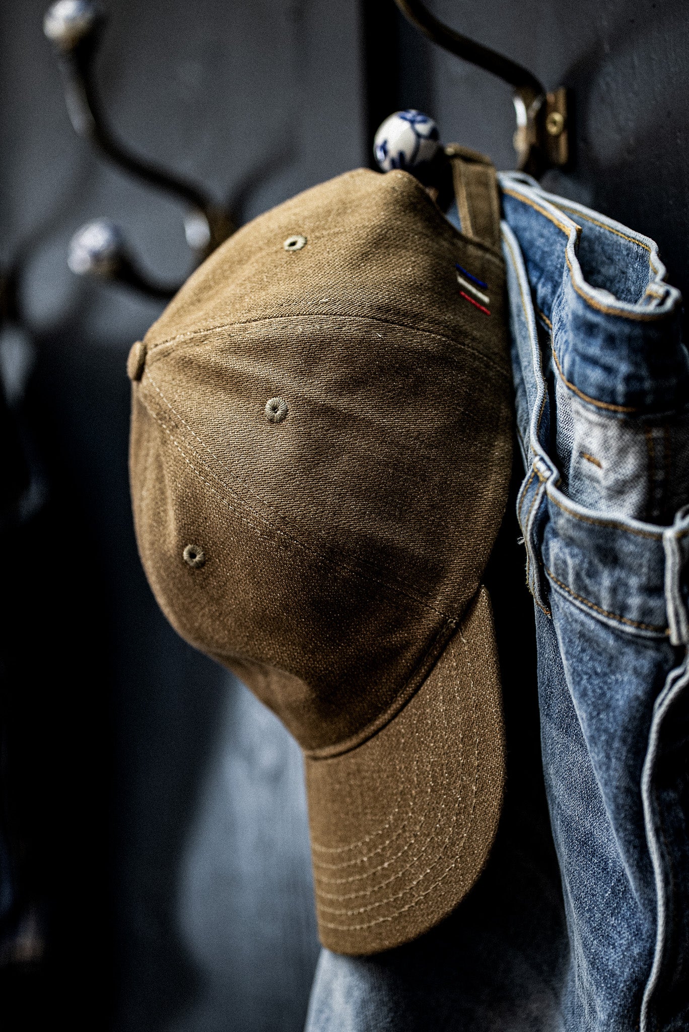 WiesMade wheat canvas ball cap hanging on a brass wall hook alongside faded denim jeans.