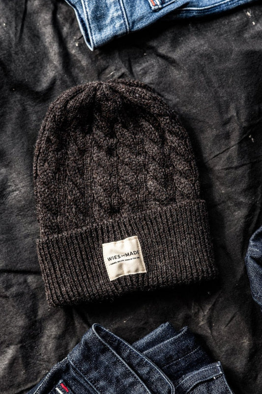 Dark basalt cable-knit wool beanie with ribbed cuff and WiesMade label, laid on black fabric beside denim.