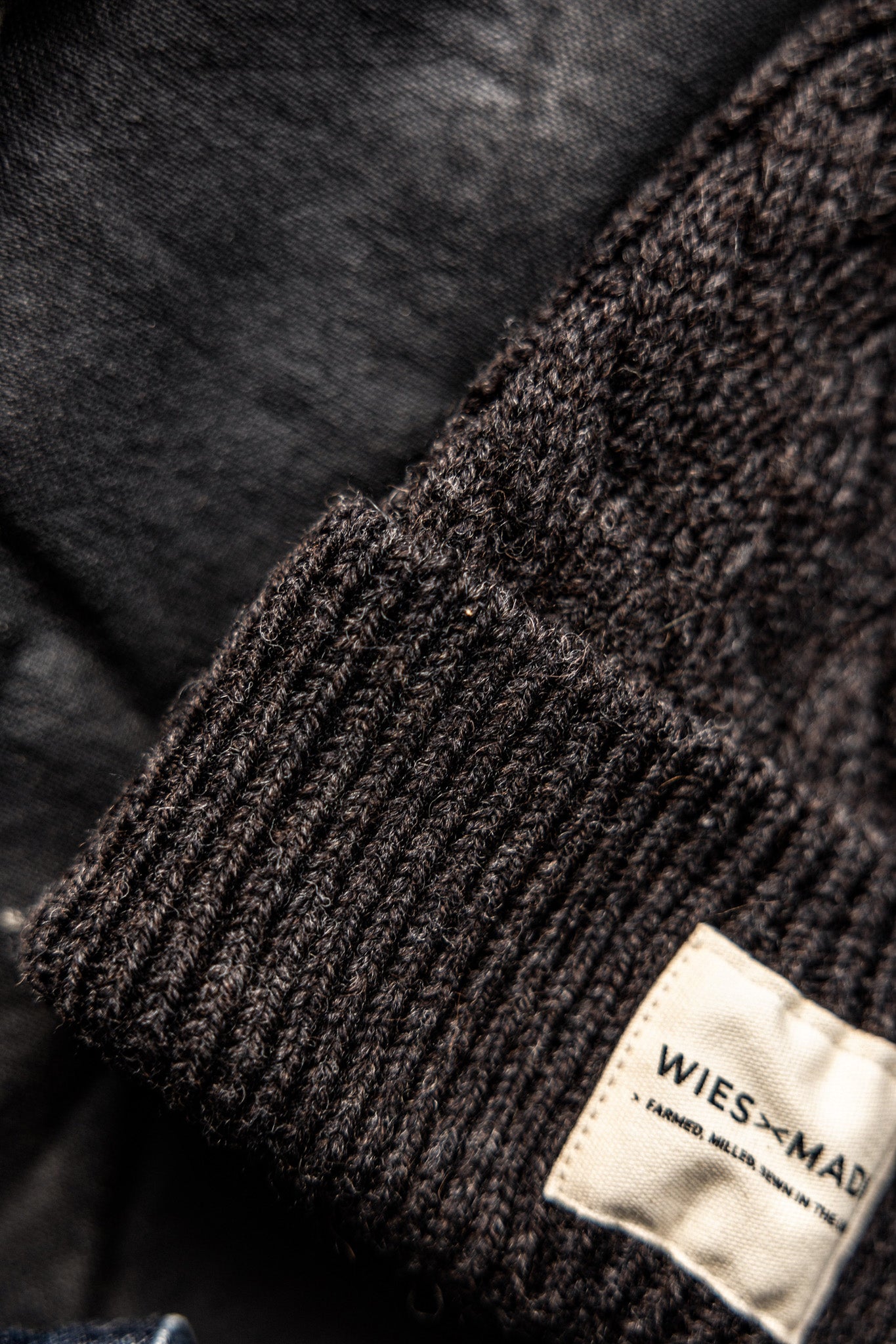 Close-up of the dark basalt wool beanie's ribbed cuff and WiesMade label on black denim.