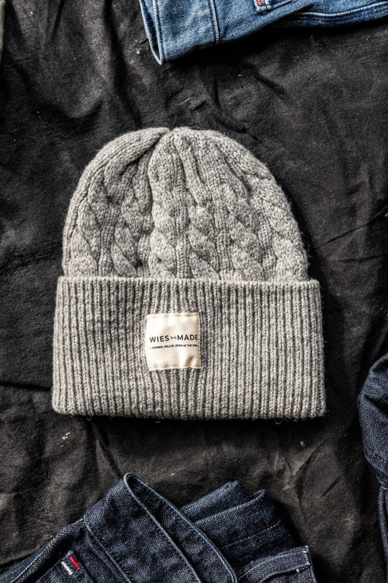 Heather grey cable-knit wool beanie with WiesMade label, laid on dark fabric between denim pieces.