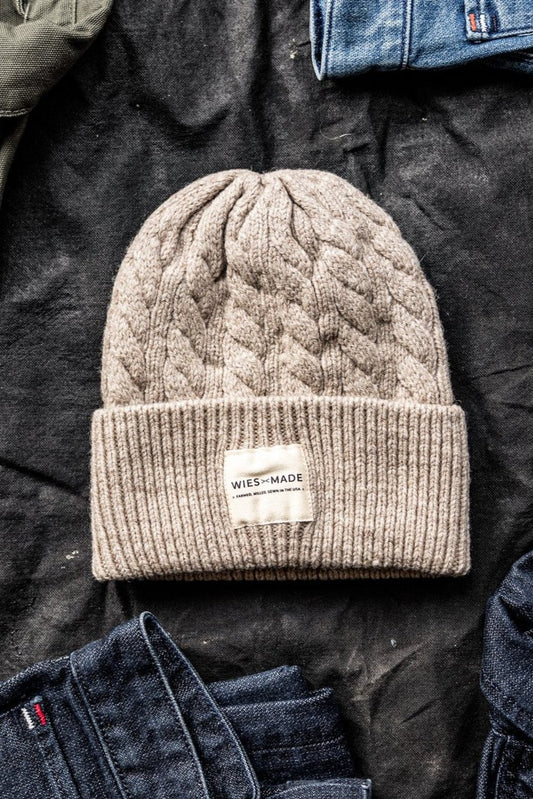 Natural cable-knit wool beanie with cuffed brim and WiesMade label, laid on dark fabric beside folded denim.