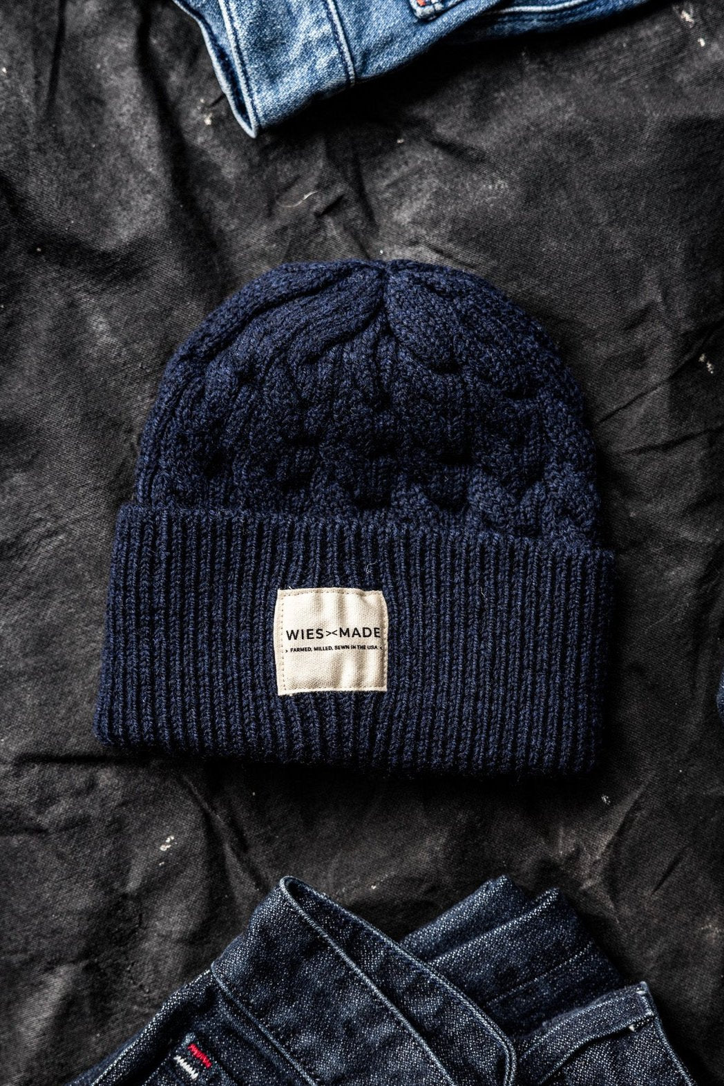 Navy cable-knit wool beanie with WiesMade label, laid on dark canvas between folded denim jeans.