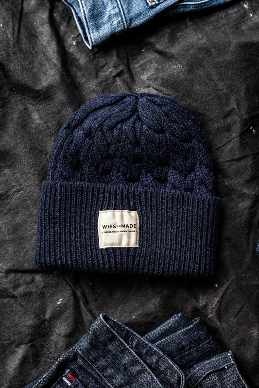 Navy cable-knit wool beanie with WiesMade label, laid on dark canvas between folded denim jeans.