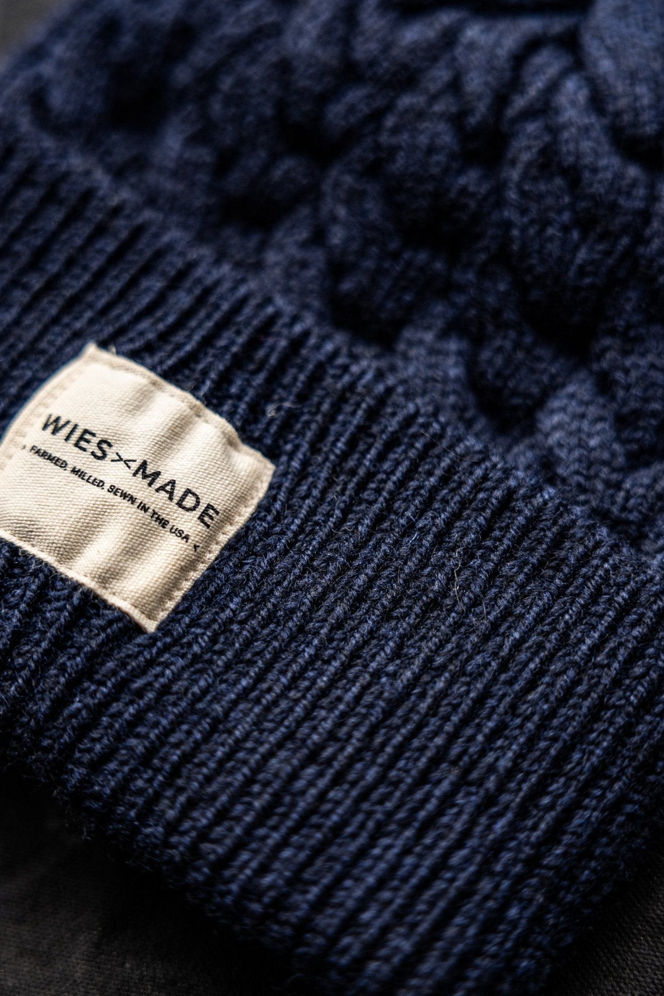 Close-up of navy cable knit wool beanie with ribbed brim and WiesMade label reading Farmed, Milled, Sewn in the USA.