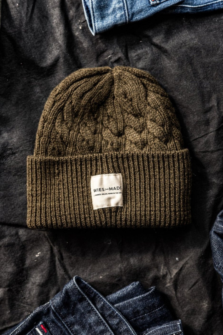 Olive cable-knit wool beanie with WiesMade label, flat-laid on dark fabric between folded denim jeans.