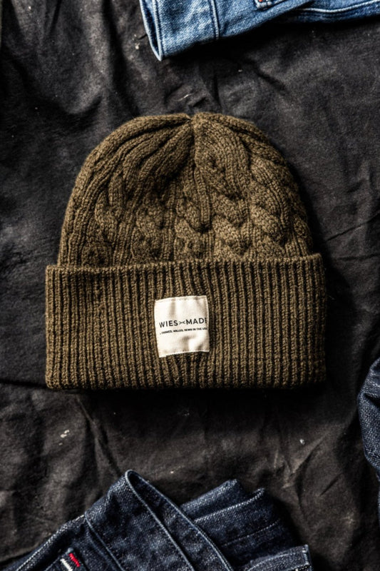Olive cable-knit wool beanie with WiesMade label, flat-laid on dark fabric between folded denim jeans.