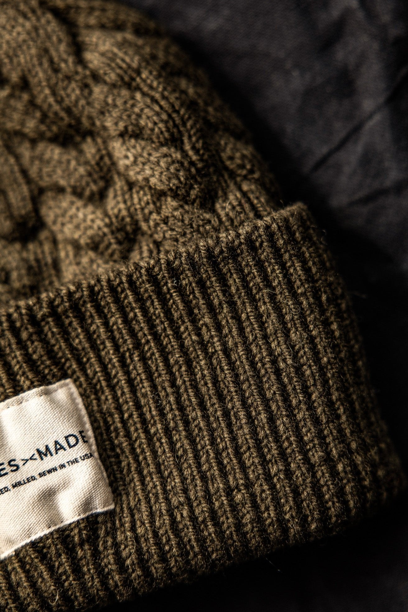 Close-up of olive cable-knit wool beanie showing ribbed cuff and WiesMade label reading Farmed, Milled, Sewn in the USA.
