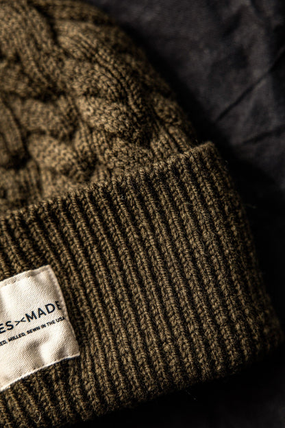 Close-up of olive cable-knit wool beanie showing ribbed cuff and WiesMade label reading Farmed, Milled, Sewn in the USA.