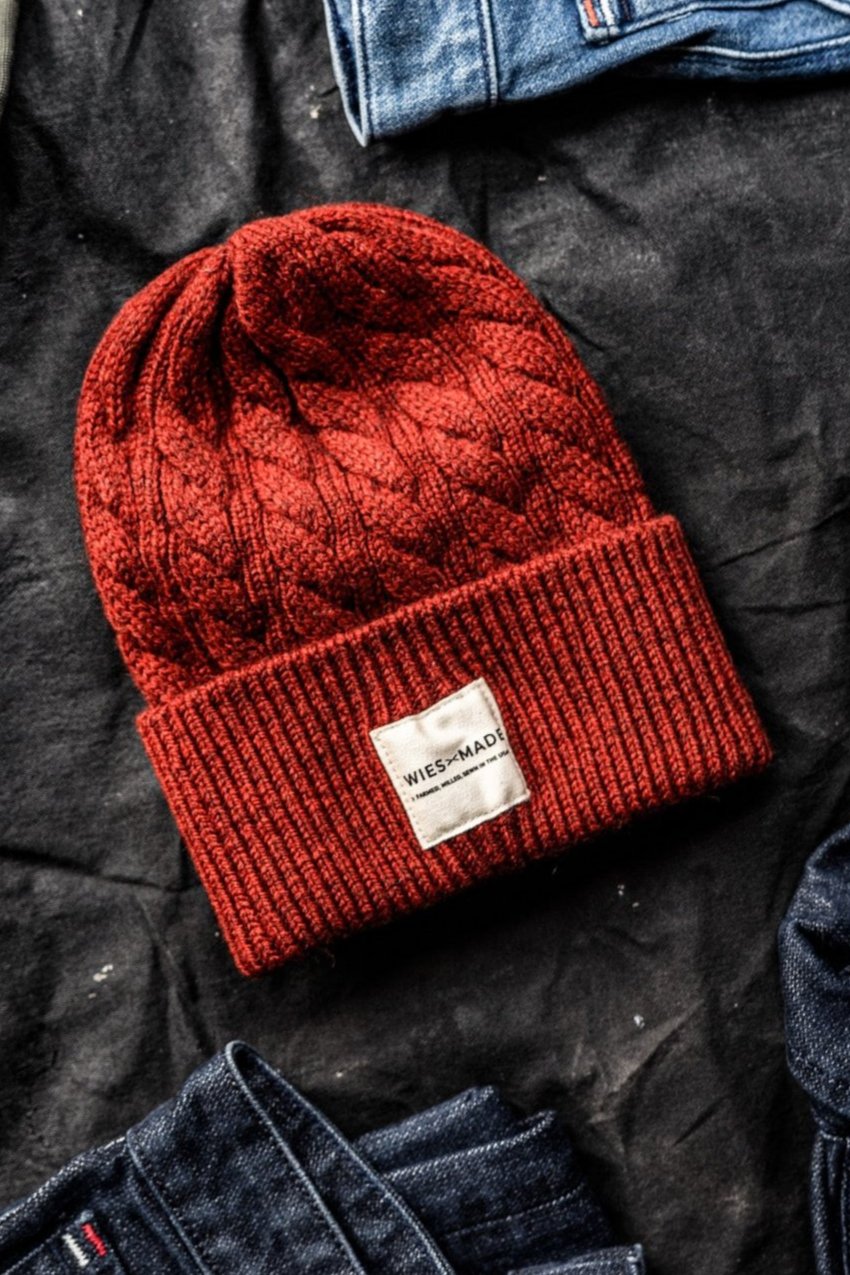Pomegranate red cable-knit wool beanie with WiesMade label, flat-lay on dark fabric beside denim jeans.