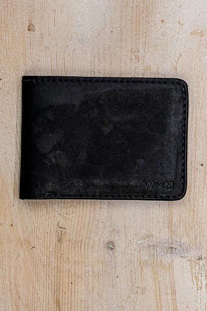 Black Horween leather bi-fold wallet with embossed WM logo on weathered wood.