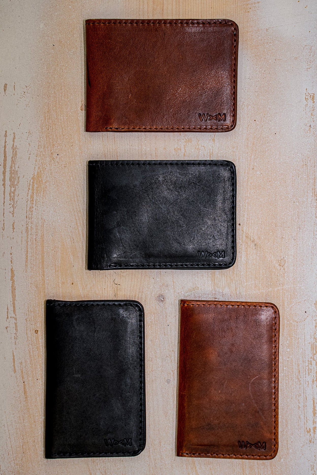 Horween leather bi-fold wallets in tan and black arranged on a weathered wood surface.