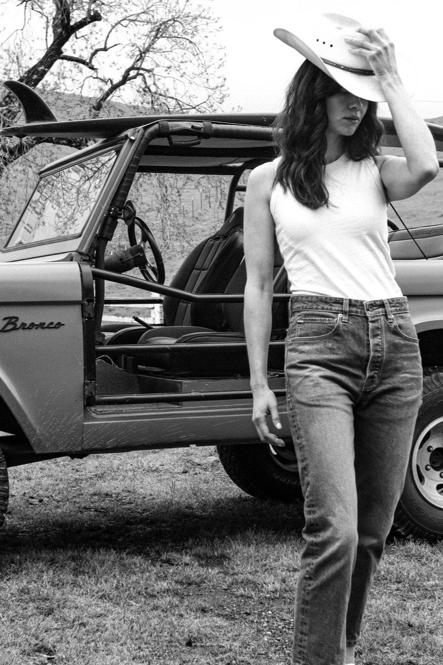 Woman in a Reyes cotton tank and jeans, wearing a cowboy hat beside a vintage Bronco.
